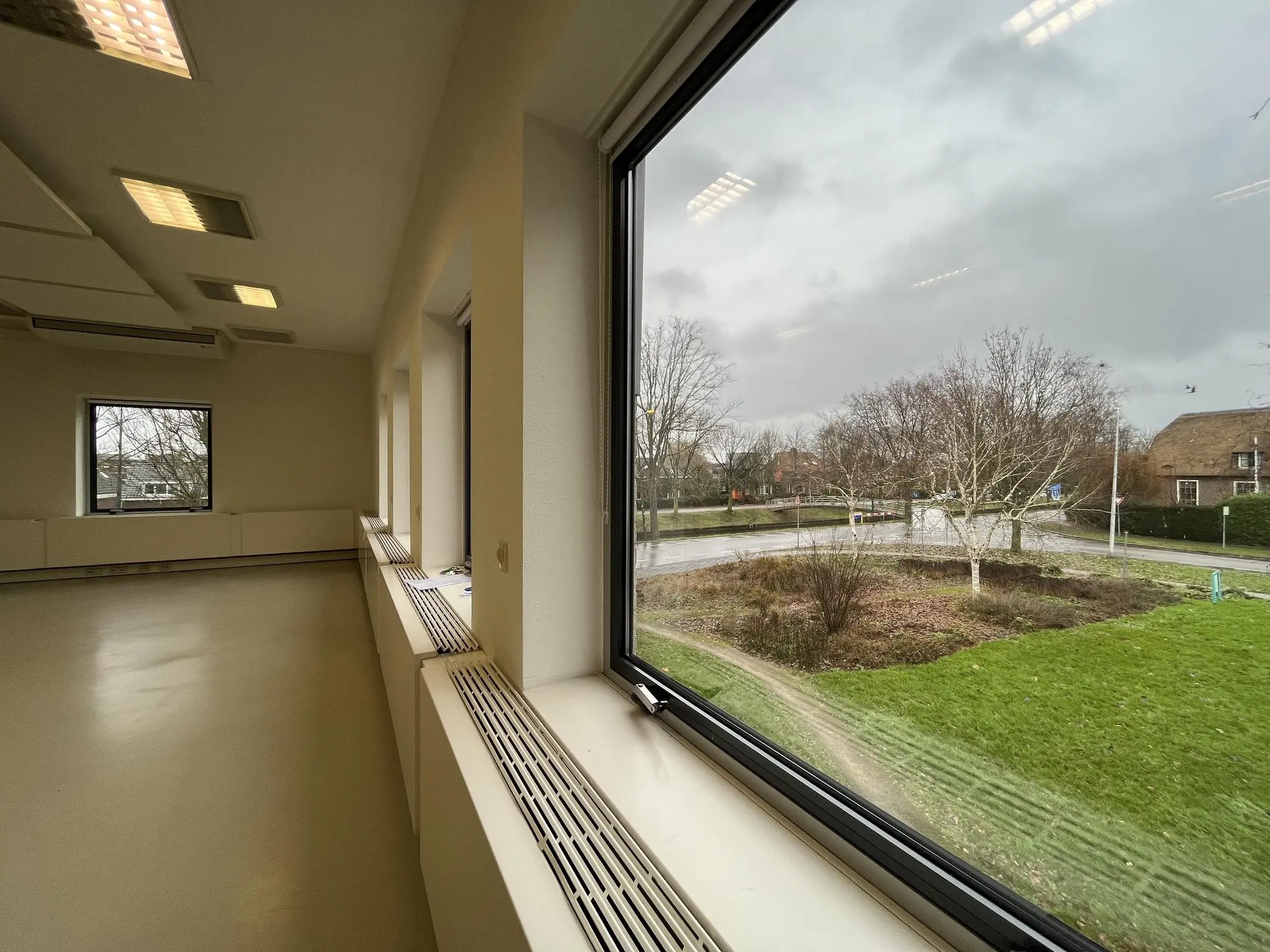 Uitzicht vanuit een leeg lokaal in Leidse Rijn op een nat en bewolkt straatbeeld met kale bomen en grasveld.